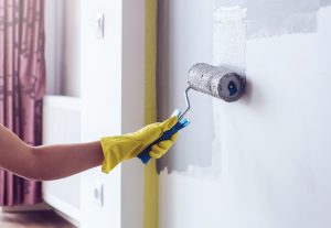 Hand in a yellow protective glove painting a wall in apartment in gray colour with a roller brush.